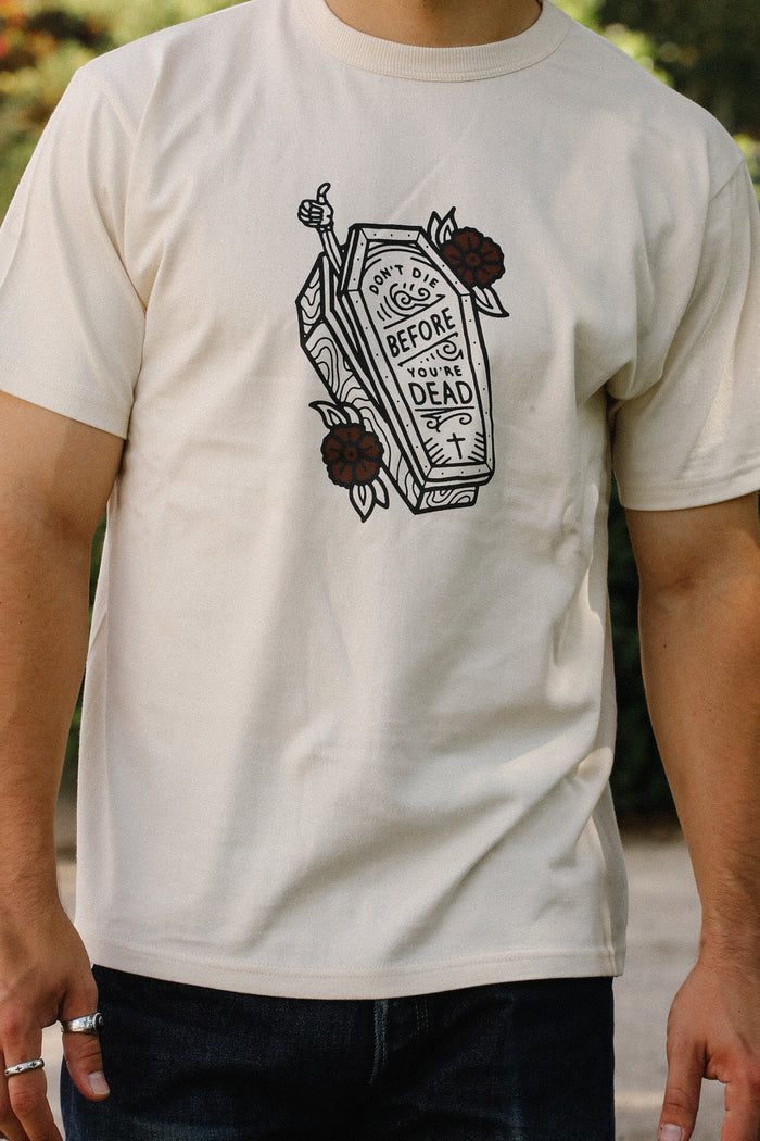 Cream Iron Shop Provisions t-shirt with coffin graphic and text 'Don't die before you're dead' worn by man outdoors