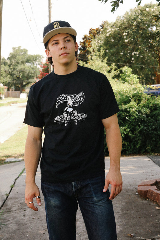 Young man wearing black loopwheel tee with vintage race car and slogan graphic, outdoors on sidewalk