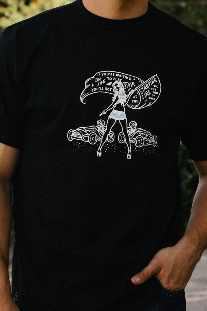 Black tee with white graphic of woman with flag and vintage race cars, motivational text about starting line and life