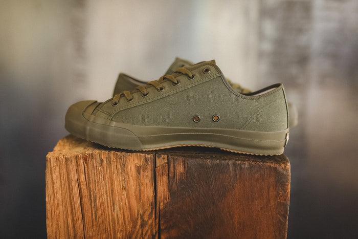 Up close image of a olive green sneaker on a wood block with a grey background