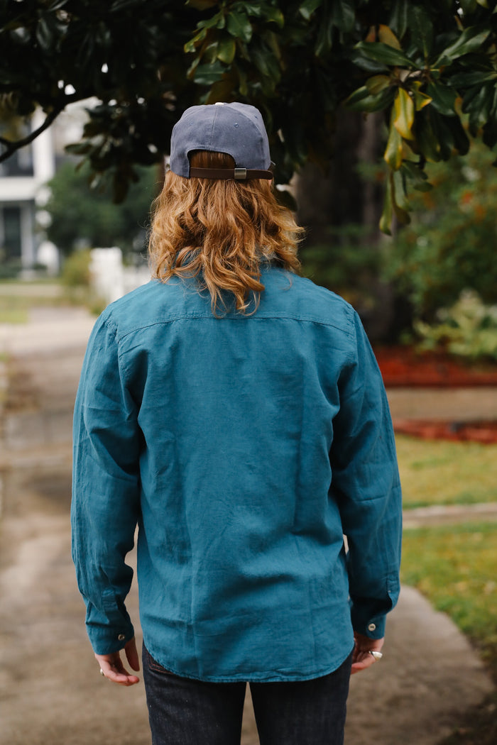 Person wearing a teal jacket and cap walking outdoors with trees and grass in the background