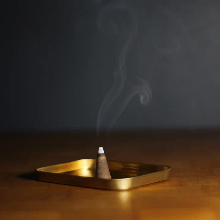 Burning incense cone emitting smoke on a gold metal tray on wooden surface