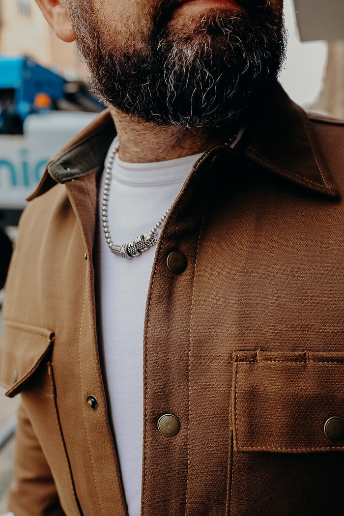 Close-up of a man with a dark beard wearing a brown button-up jacket and a silver beaded necklace over a white shirt