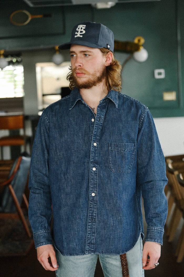 Man wearing navy blue denim shirt and dark baseball cap indoors in casual setting