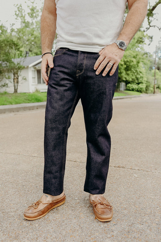 Man wearing dark raw denim jeans, white t-shirt, brown leather moccasins, standing outdoors on street