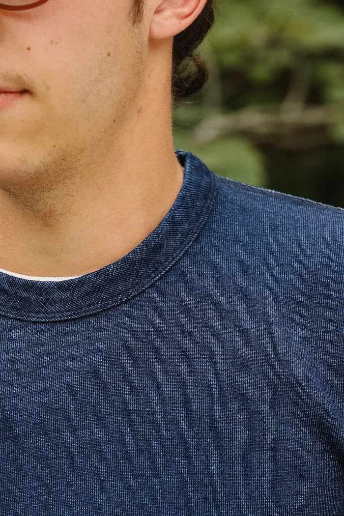 Close-up of a person wearing a dark navy blue crewneck knit shirt, showing the collar and textured fabric against a blurred green outdoor background.