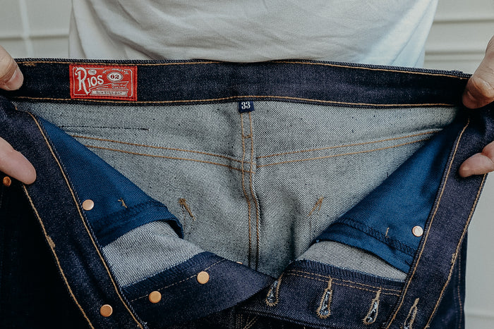 Hands holding open dark blue Rios slim straight denim jeans showing inner stitching and size 33 tag