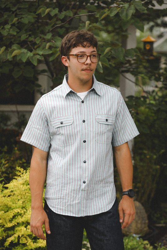 Person wearing a striped shirt and glasses standing outdoors with greenery in the background