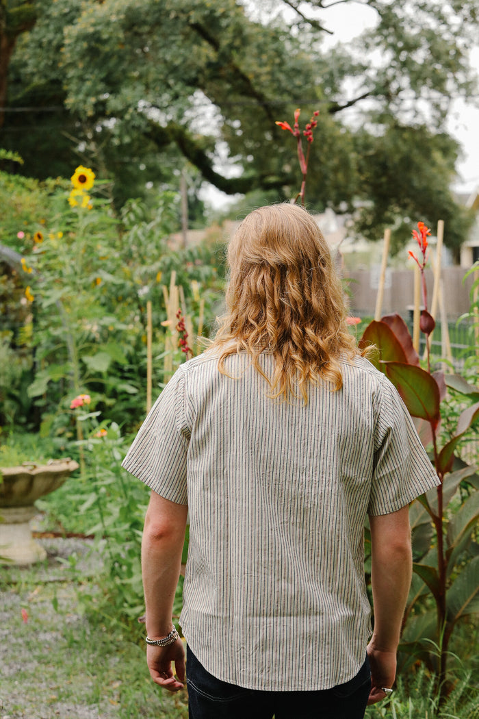 Person with long hair standing in a garden with various plants and flowers.