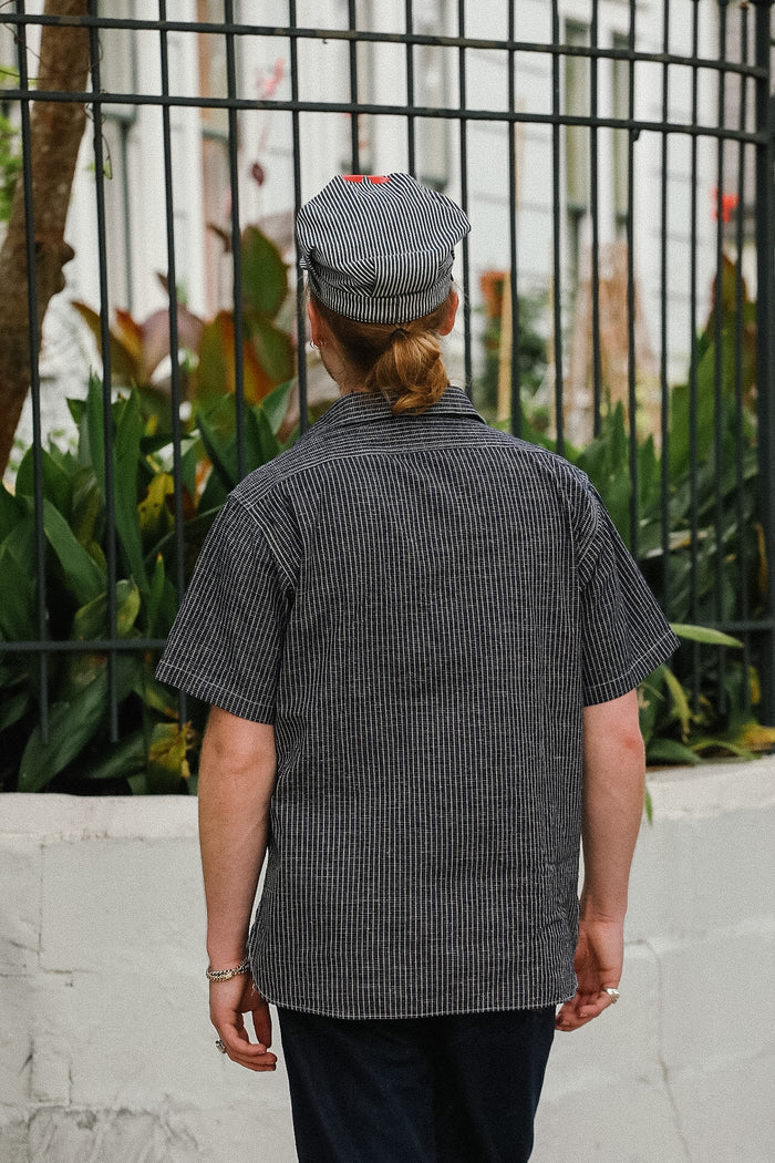Person wearing a black and white checkered shirt with a cap, standing in front of a metal fence and plants.