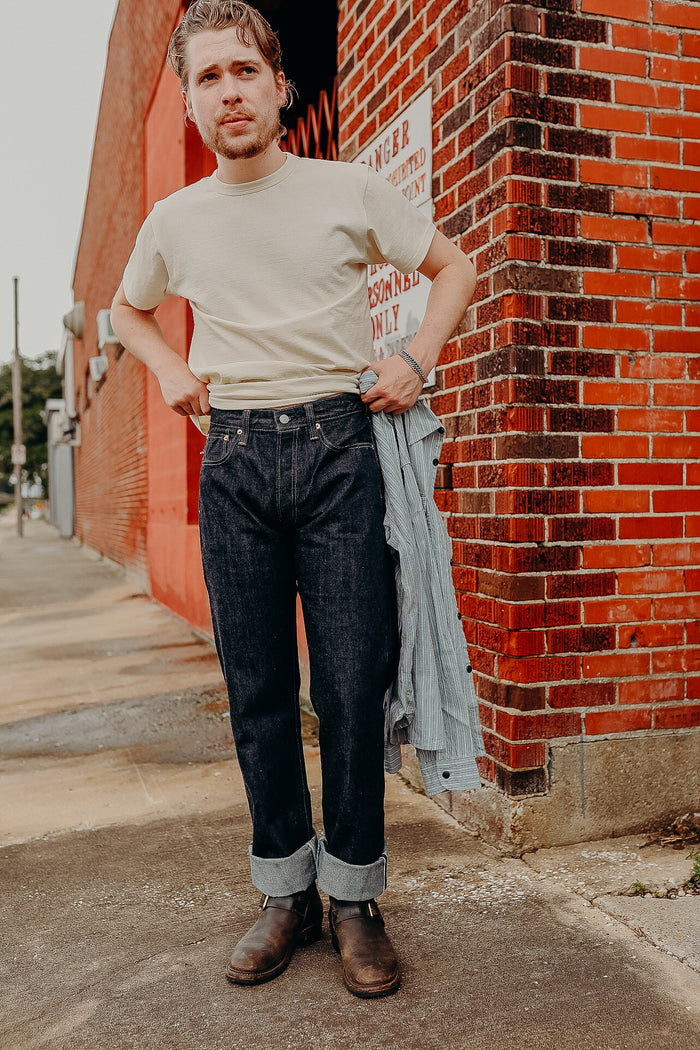 Man wearing beige t-shirt, dark cuffed jeans, and brown leather boots standing by red brick wall holding striped shirt