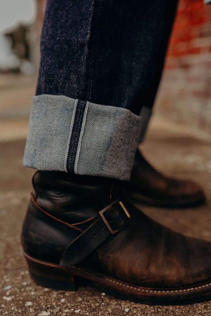 Close-up of cuffed dark denim jeans paired with worn brown leather engineer boots on concrete floor