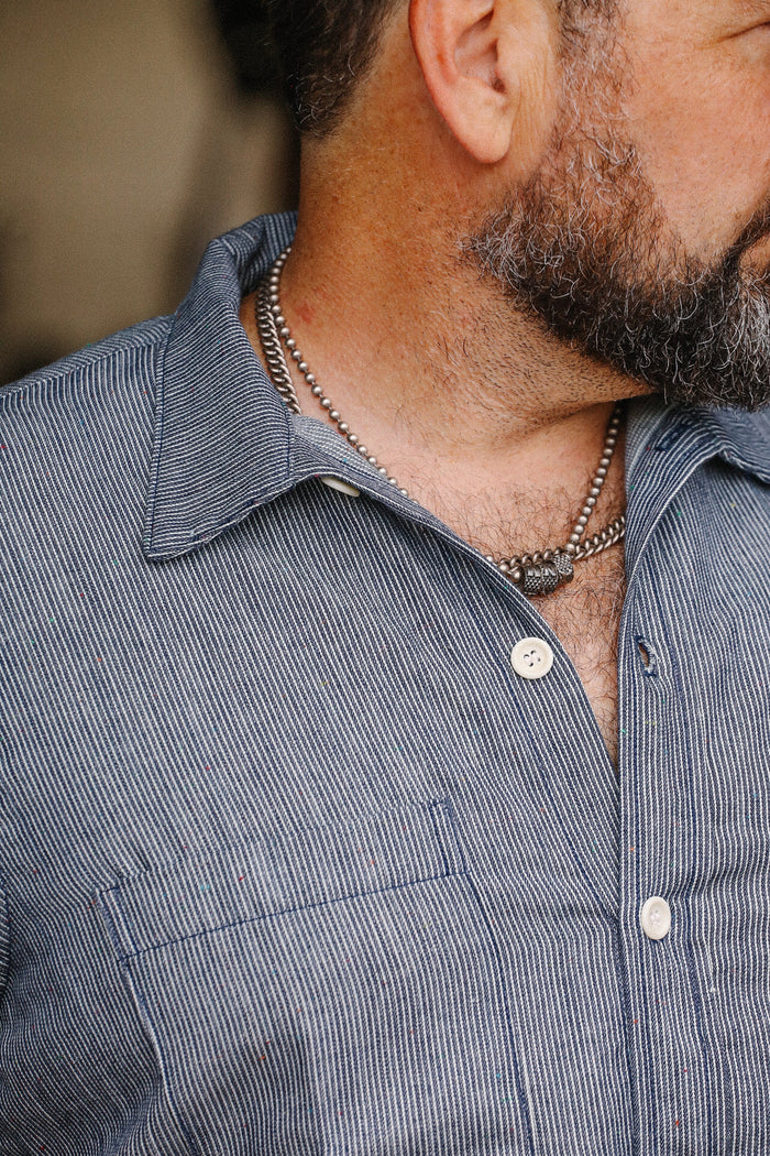 Close-up of a man wearing a blue striped v-short sleeve shop shirt with white buttons and a silver chain necklace