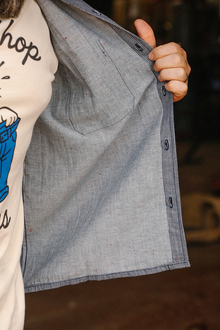Close-up of a hand holding open a blue and white striped shirt with rainbow nep details inside, worn over a graphic tee at Iron Shop Provisions