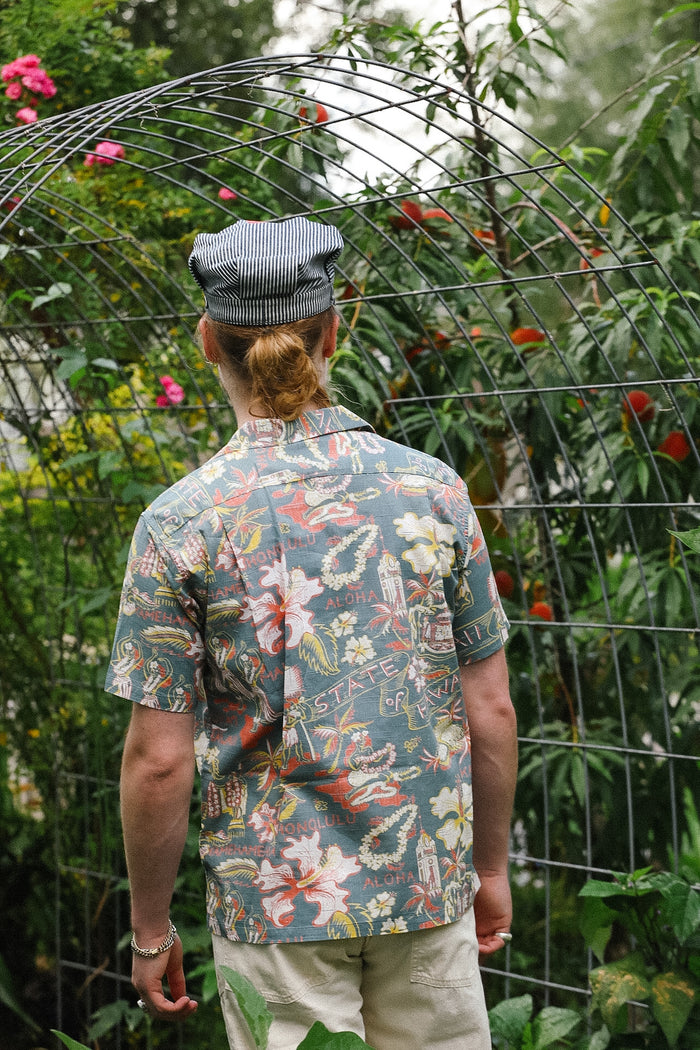Person wearing a patterned shirt and cap standing in front of a wire arch with plants