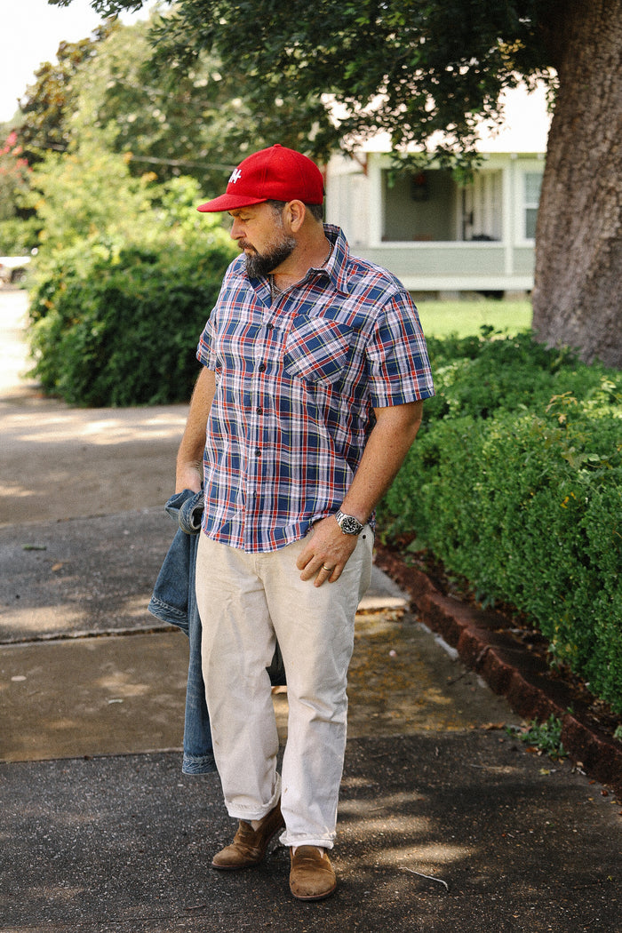 Man wearing navy red plaid short sleeve madras shirt, beige pants, brown shoes, and red cap outdoors