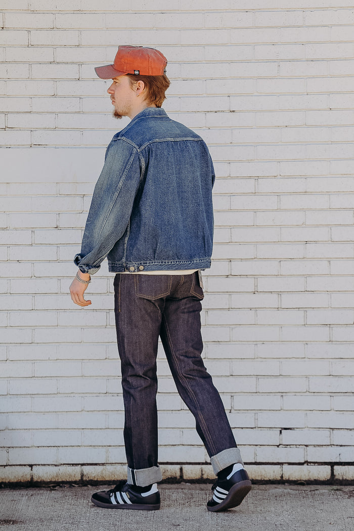 Man in blue denim jacket, dark jeans, and orange cap walking against white brick wall