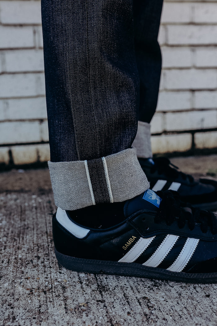 Close-up of rolled raw denim jeans paired with black Adidas Samba sneakers on textured concrete floor