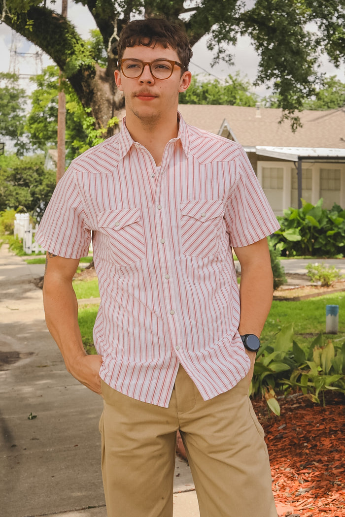 Man wearing a red and white striped shirt and beige pants standing outdoors.