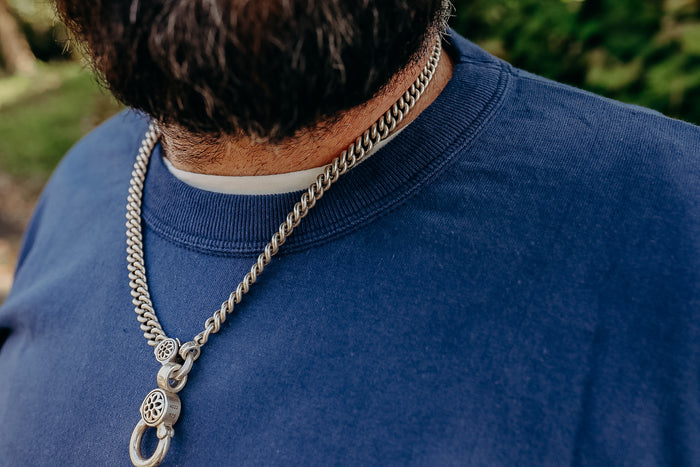 Close-up of a man wearing a silver chain necklace with detailed clasps over a blue crewneck shirt