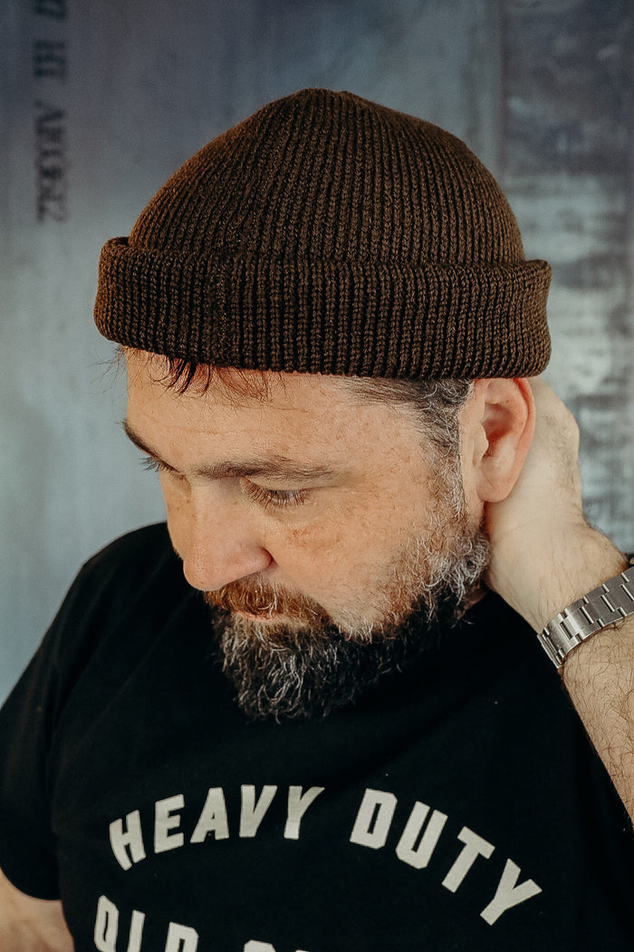 Man wearing brown knit beanie and black heavy duty t-shirt, looking down with hand behind neck