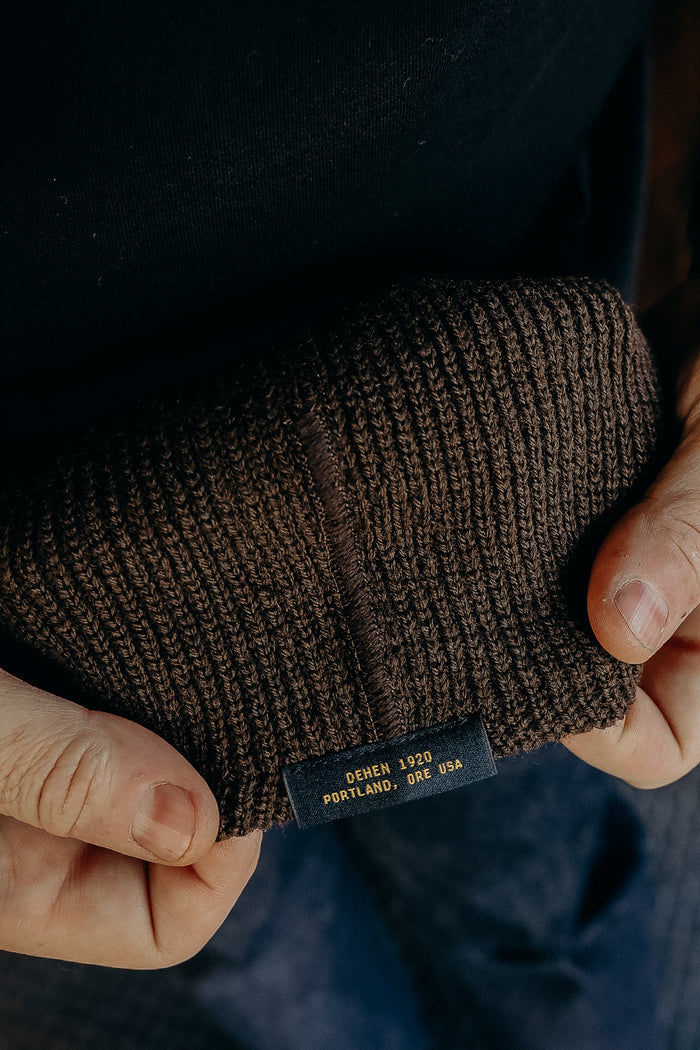 Close-up of hands holding a brown knitted beanie with Dehen 1920 Portland, Ore USA label