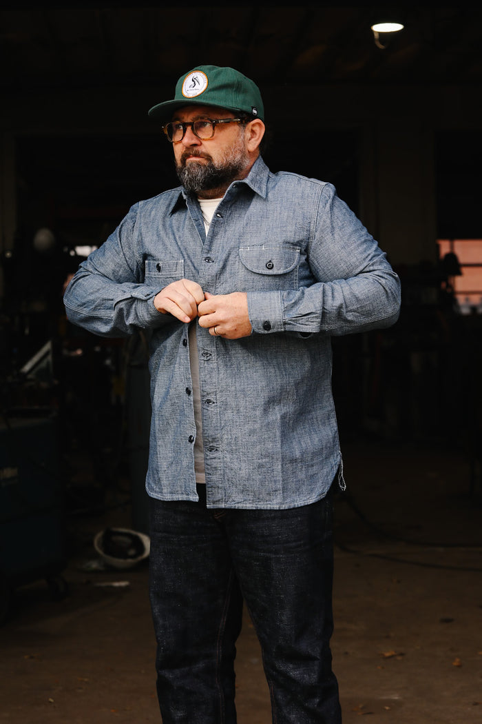 Man wearing green cap and blue chambray work shirt buttoning up in industrial workshop