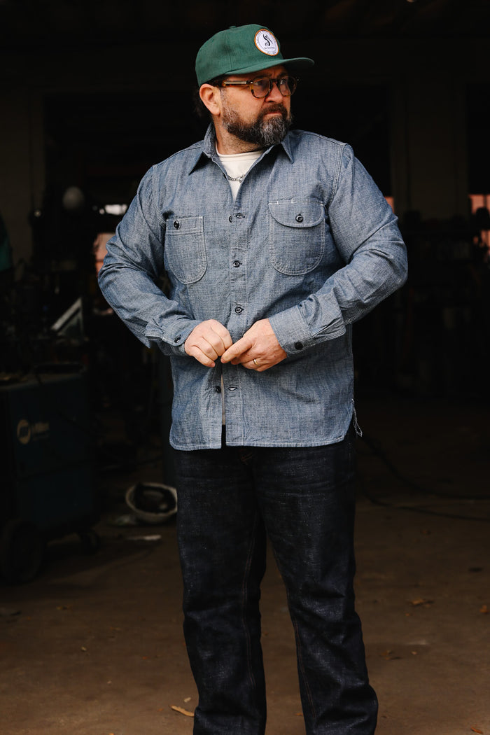 Man wearing denim work shirt, dark jeans, green cap, and glasses in workshop setting