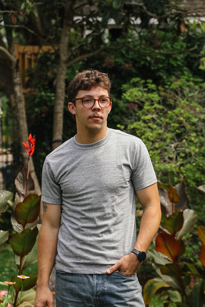 Man wearing a gray t-shirt and glasses standing in a garden with greenery.