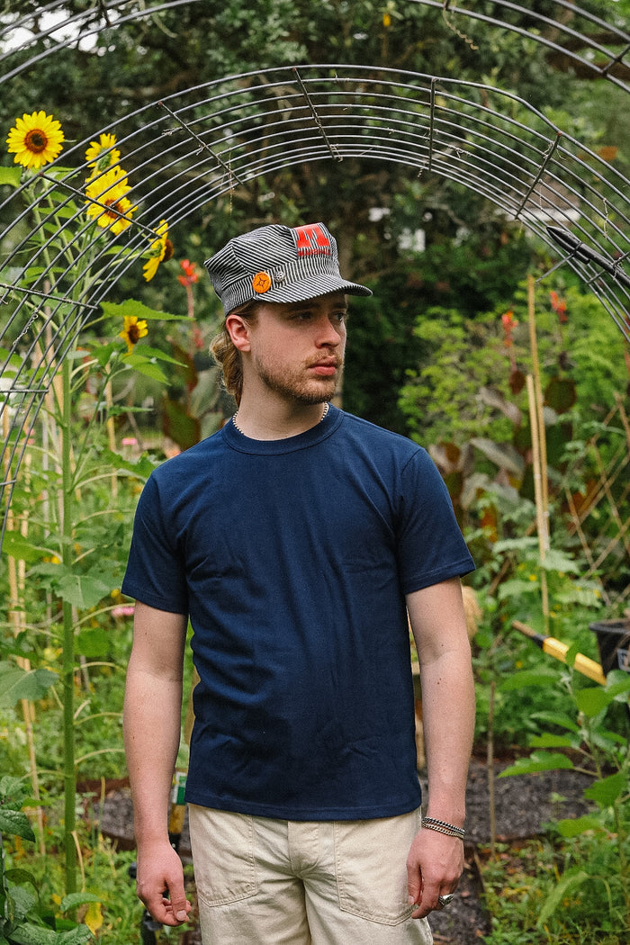 Man wearing a cap and blue shirt standing in a garden with sunflowers and greenery.